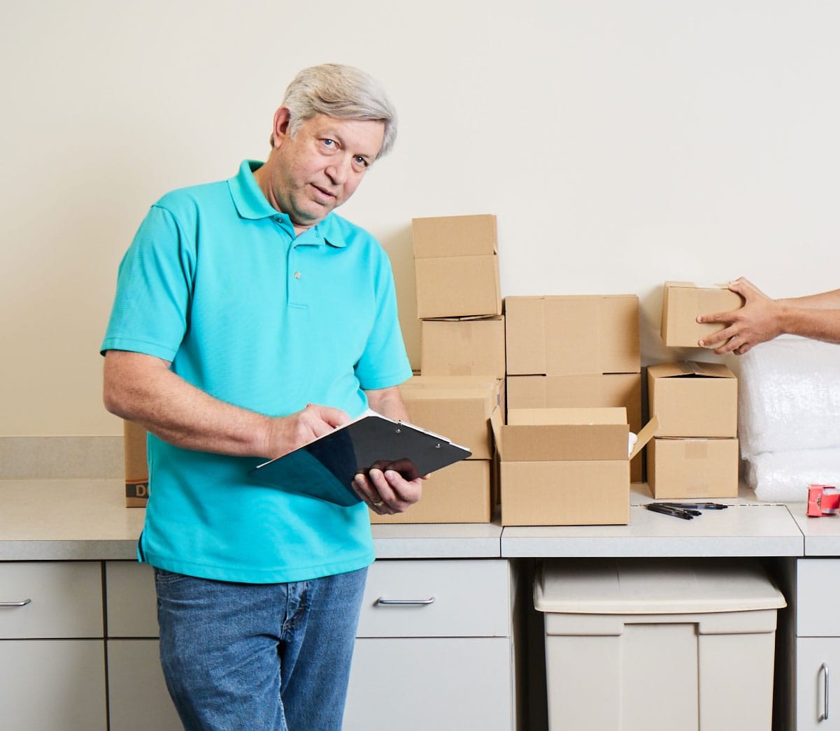An older man with gray hair in a turquoise polo shirt and jeans stands in front of a counter with stacked cardboard boxes. He holds a clipboard and appears to be taking notes. Another hand reaches to stack a box.