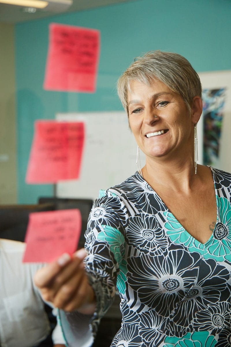 A smiling person with short gray hair places pink sticky notes on a glass surface in an office setting. They are wearing a patterned dress with floral designs. In the background, there's a whiteboard and a blurred office environment.