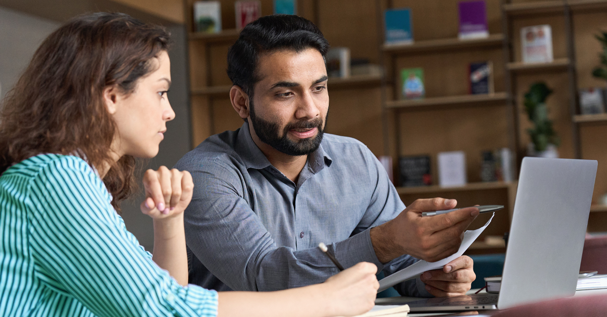 A man and woman sit at a table with a laptop, engaged in discussion. The man points at the laptop screen while holding papers, and the woman listens attentively with a pen in hand. In the background, books are displayed on shelves.