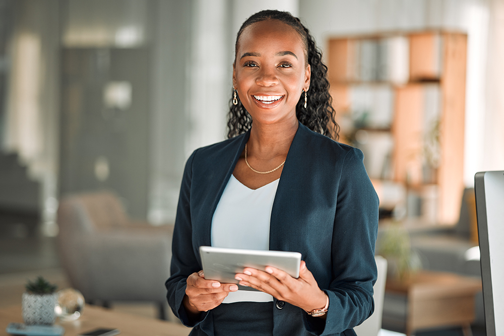 A smiling woman in a business suit holds a tablet in a modern office setting, reviewing payment terms. She stands in front of blurred shelves and furniture, exuding a professional and confident demeanor.