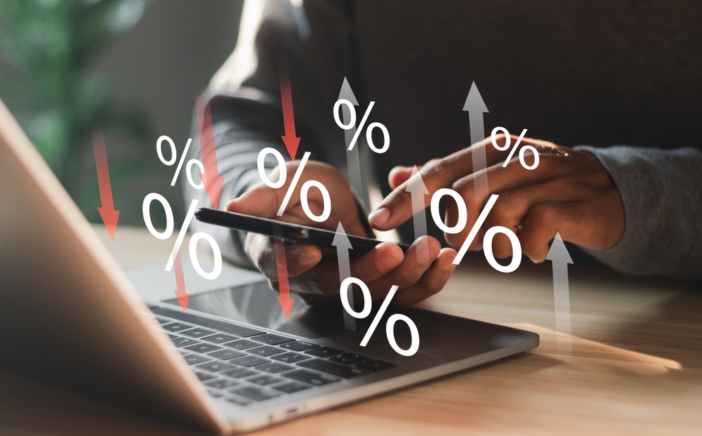 A person using a smartphone and laptop with percentage symbols and arrows pointing up and down, representing fluctuating interest rates, early payment strategies, or financial data trends affecting cash flow.
